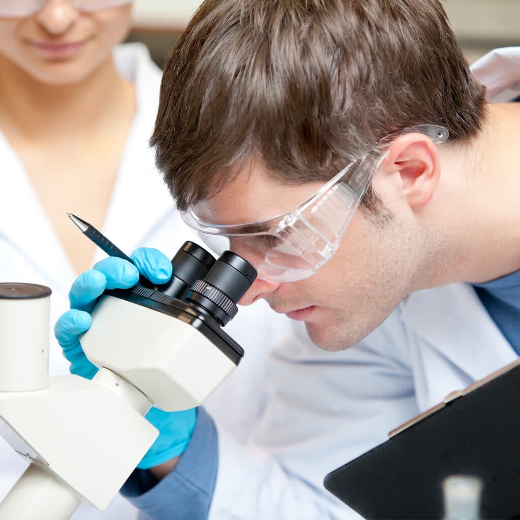 Testimonials from contract testing services providers. Medical researcher smiling while looking through a microscope for a microbiology testing