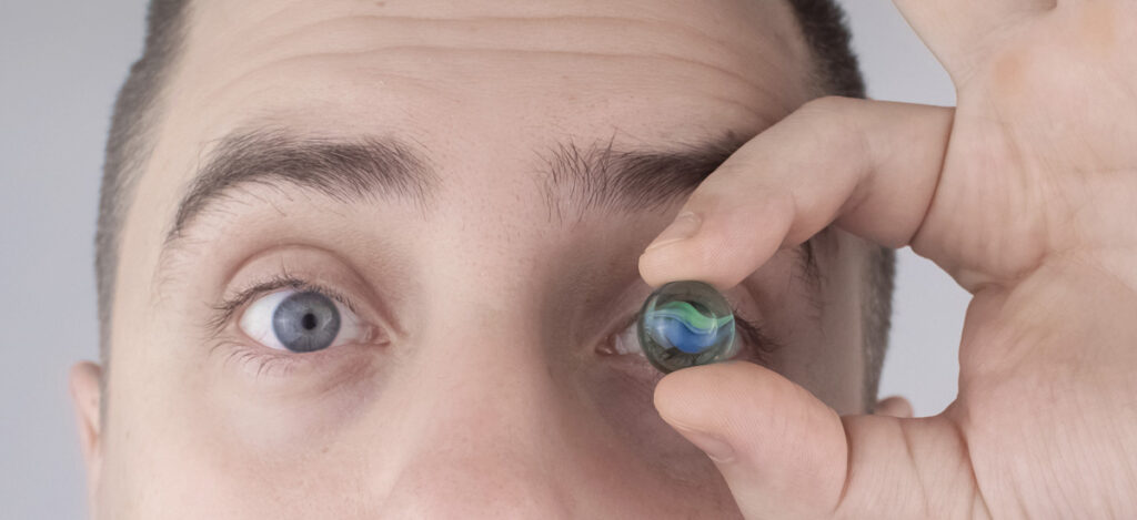Close frontal picture of a person holding with his fingers an eye implant in front of one of his eyes. Biocompatibility tests for medical devices. Biocompatibility tests for medical implants. Medical implants biocompatibility tests. Medical devices biocompatibility tests. What is compatibility testing. Compatibility testing definition. USP guidance for biocompatibility tests. Biocompatibility tests regulatory requirements. Medical devices categorization for biocompatibility testing. How do you test biocompatibility of medical devices